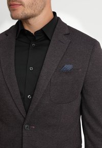Blazer a spina di pesce scuro in un tessuto texturizzato, con revers a punta, taschino sul petto con un fazzoletto blu a pois e una camicia nera sottostante.