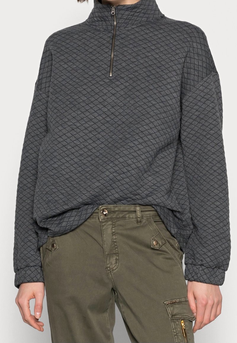 Grijze gewatteerde pullover met een hoge kraag en een halve rits. Draag met olijfgroene cargobroek met zakken en gouden accenten.