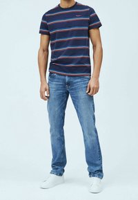 Camisa de rayas azul marino con rayas rojas y azules, combinada con jeans de color azul claro y zapatillas blancas. Ajuste casual, tejido de algodón.