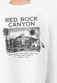 Biela košeľa s dlhým rukávom zobrazuje čiernu grafiku kovboja na koni, obklopeného prvkami púšte a textom "RED ROCK CANYON."