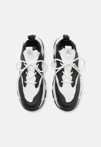 Zapatillas deportivas en blanco y negro con malla texturizada y paneles de cuero liso, que cuentan con punta redondeada, diseño con cordones y suela gruesa.