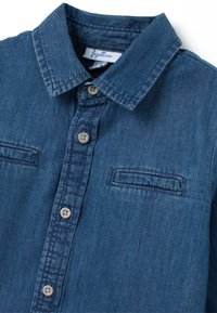 Camisa de mezclilla en azul oscuro con cuello, dos bolsillos en el pecho y botones plateados. Textura suave y diseño estructurado.