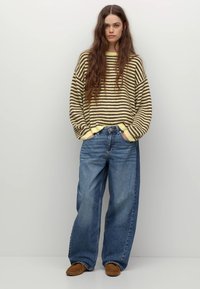Pull en laine rayé jaune et marron, associé à un jean en denim bleu ample. Le mannequin se tient sur un fond neutre, portant des chaussons marron.