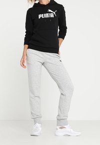 Sweat à capuche noir avec logo "PUMA" blanc, assorti à un pantalon de survêtement gris clair. Le modèle porte des chaussures de sport blanches avec des accents gris.