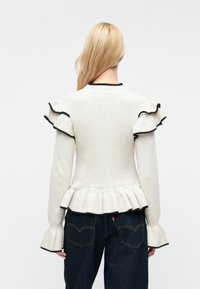 Rebrast pulover v kremni barvi z črnimi detajli, nagubanimi rokavi in peplum robom. Ima mehak usnjeni material in prilegajočo obliko, v kombinaciji s temnimi džinsom.