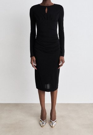 Black dress with long sleeves, ruched detailing, and a front keyhole. Worn with silver and gold accent high heels. Smooth texture.