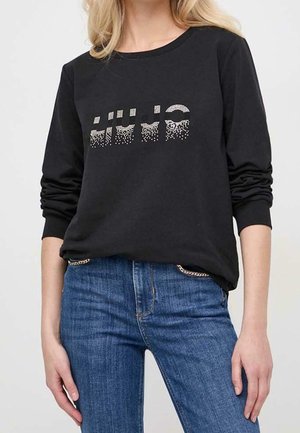 Mujer que lleva una camiseta negra de manga larga con puntos brillantes que forman un patrón abstracto y vaqueros azules con detalles de cadenas en los bolsillos.