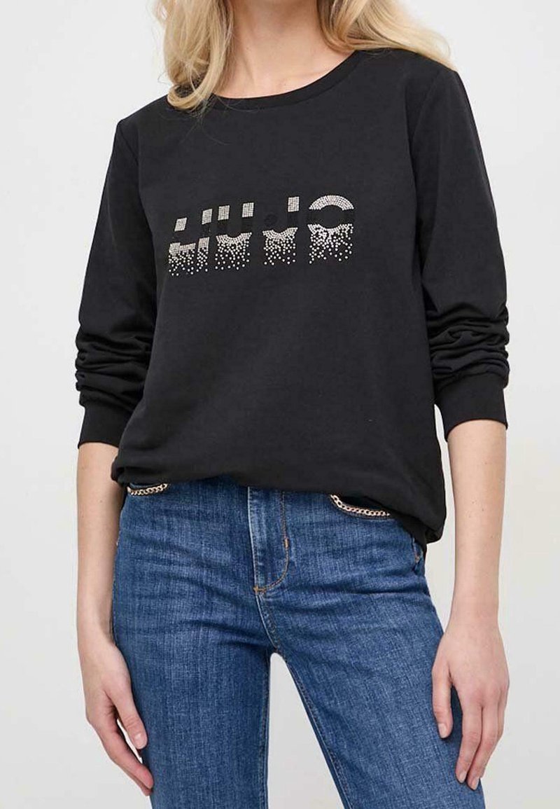 Femme portant un haut noir à manches longues avec des points scintillants formant un motif abstrait et un jean bleu avec des détails en chaîne sur les poches.