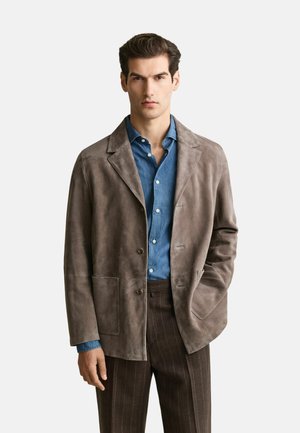 Man wearing a gray suede jacket over a blue button-up shirt and brown pinstripe trousers, standing with one hand in his pocket.