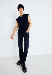 Top in maglia senza maniche di colore blu navy abbinato a jeans di denim scuro e scarpe sportive nere con accenti verdi. Il modello sta in piedi con sicurezza con le mani in tasca.