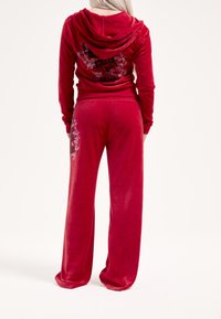 Červená velúrová tepláková súprava obsahujúca mikinu s kapucňou a grafickým textom a rozšírené nohavice s ozdobnou potlačou na boku. Mäkká textúra, voľný strih.