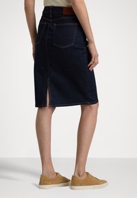 Femme portant une jupe en denim bleu foncé, longueur genoux, avec une fente à l'arrière, un haut beige et des baskets en daim tan, se tenant sur un sol blanc.