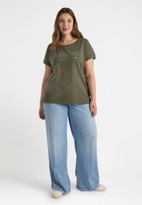 Olivgrön t-shirt med rundad halsringning, prydd med ett tryckt slogan. Kombineras med vida ljusblå jeans och beiga sneakers.