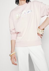 Svetle růžová mikina s uvolněným střihem. Na přední straně je bílý a duhový nápis "Hugo 1993". Kombinováno s bílými kalhotami a naskládanými náramky.