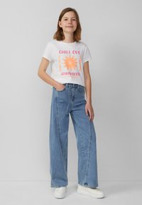 T-shirt blanche à manches courtes avec un graphique de soleil orange et du texte, associée à un jean large bleu clair. Accessoirisée avec des baskets blanches.