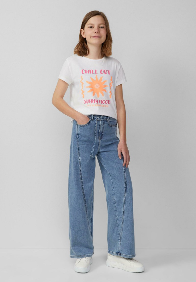 T-shirt blanche à manches courtes avec un graphique de soleil orange et du texte, associée à un jean large bleu clair. Accessoirisée avec des baskets blanches.