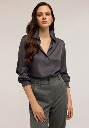 Donna che indossa una blusa in satin grigio scuro con colletto rialzato e pantaloni a righe grigi a vita alta, in piedi su uno sfondo neutro.