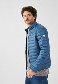 Giacca trapuntata blu con colletto alla coreana e logo sulla manica sinistra; indossata sopra un maglione a motivo e jeans di denim.