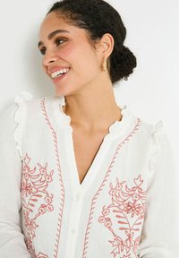 Camisa de lino blanca con bordados florales en tonos rosas, cuello con volantes y cierre de botones. Textura suave con costuras y detalles elaborados.
