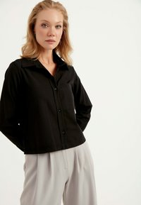 Svart långärmad skjorta med krage, framficka och knäppning. Modellen bär ljusgrå byxor som visar en avslappnad passform.