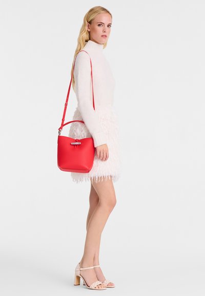 Red bucket bag with a smooth texture and silver hardware. Adjustable shoulder strap. Worn with a light pink fluffy dress and beige heels.