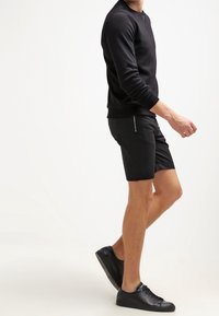 Schwarzer Crewneck-Sweatshirt und schwarze Shorts aus glattem Stoff. Die Shorts haben einen Reißverschlussakzent an der Seite und werden mit schwarzen Turnschuhen kombiniert.