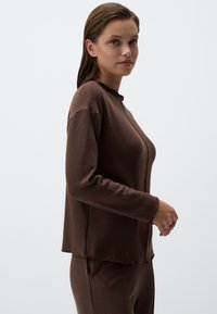 Top lungo a maniche lunghe di colore marrone con scollatura rotonda e orlo irregolare. Il tessuto sembra morbido e leggermente elasticizzato. Modello in posa di lato.