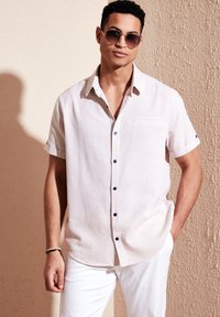 Camisa de lino de manga corta en color beige claro con una textura sutil, con cuello abotonado y botones azul marino. Pantalones blancos y gafas de sol.