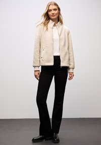 Cremefarbene Fleecejacke mit hohem Kragen, Reißverschluss vorne und seitlichen Taschen. Dazu schwarze Hosen und Stiefeletten.