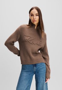 Braun geripptes Strickoberteil mit hohem Kragen und besticktem "Love"-Detail. Kombiniert mit blauen weiten Jeans, die lange Ärmel haben.