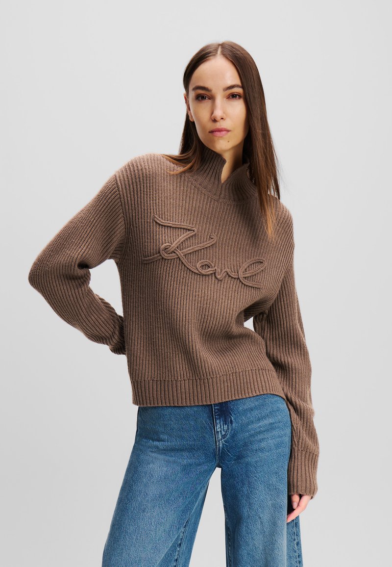 Braun geripptes Strickoberteil mit hohem Kragen und besticktem "Love"-Detail. Kombiniert mit blauen weiten Jeans, die lange Ärmel haben.