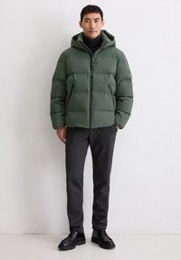 Veste doudoune verte avec capuche, fermeture éclair et poches latérales. Portée sur un col roulé noir, associée à un pantalon sombre et des chaussures noires.