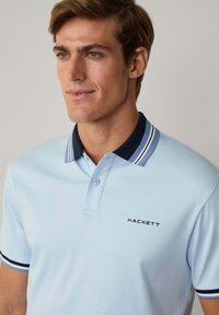 Světle modrá polo košile s límcem pruhovaným v námořnické modré a bílé barvě, dvouknoflíkovou légou a černým logem "HACKETT" na hrudi. Hladká textura.