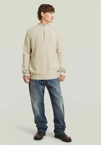 Beige trui met ritssluiting en gegolfde ribbels, gecombineerd met lichtblauwe jeans en donkerbruine laarzen. Eenvoudig ontwerp, comfortabele pasvorm, geen patronen.