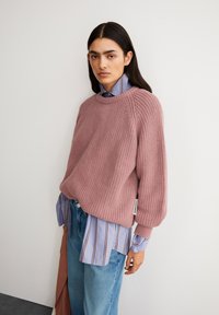 Rosa gestrickter Pullover mit geripptem Muster, Rundhalskragen und落降te Schultern, übereinander getragen mit einem hellblauen gestreiften Hemd und Jeans.