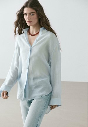 Femme aux longs cheveux bruns portant une chemise à boutons bleu clair, un jean en denim clair, et un collier de perles rouges, marchant à l'intérieur.