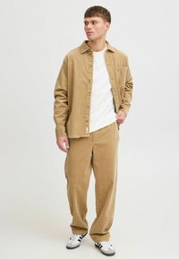 Hellbeige Kordjacke mit Knopfverschluss und Brusttasche, getragen über einem weißen Rundhals-Shirt und passenden locker sitzenden Hosen.