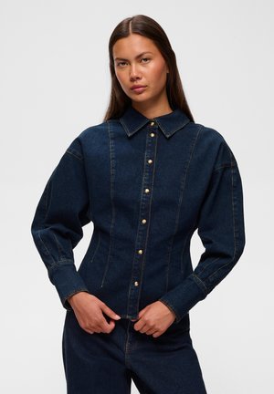 Femme aux longs cheveux foncés portant une veste en denim foncé ajustée avec des boutons dorés et un pantalon en denim foncé assorti sur un fond uni.