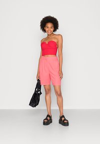 Top sem alças vermelho com detalhe de volantes, calções rosa ajustados e sandálias de plataforma pretas. Modelo segura uma bolsa preta com alças em nó.