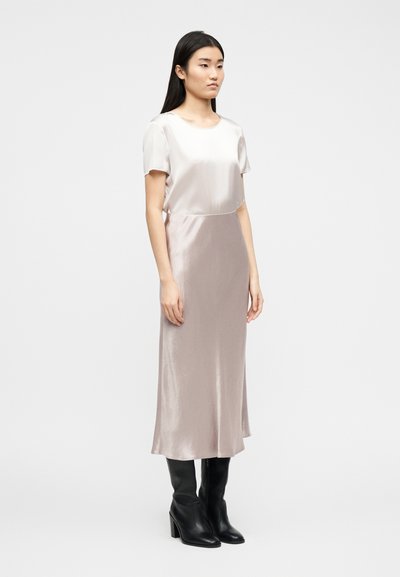 Γ υναίκα με μεταξωτό μπλουζάκι με κοντά μανίκια και μακριά φούστα στο ίδιο χρώμα, φοράει μαύρες μπότες με τακούνια, στέκεται μπροστά σε απλό λευκό φόντο.