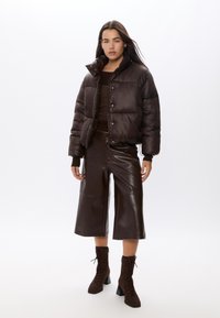 Giacca a vento marrone con colletto alto, texture lucida e bottoni a scatto, abbinata a culotte in pelle marrone e stivaletti coordinati.