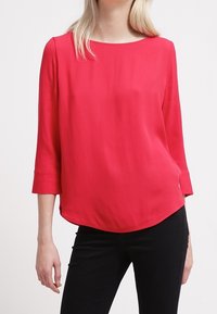 Blusa ligera roja con cuello redondo, mangas de tres cuartos y dobladillo curvado. Textura suave y sin patrones o accesorios visibles.