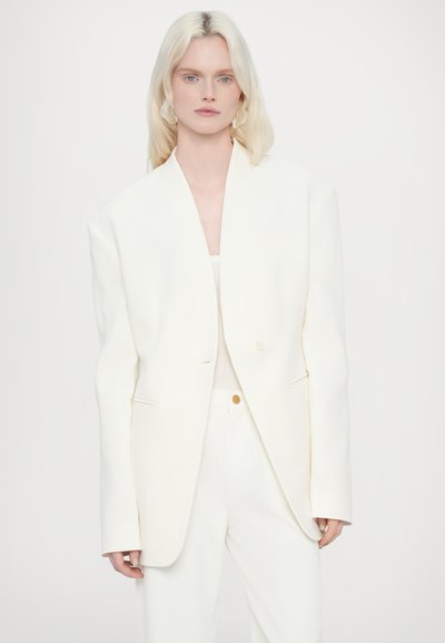 Witte oversized blazer met een diepe v-halslijn, gestructureerde schouders en klepzakken, gecombineerd met witte broek en een lichte top.