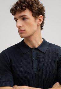 Donkerblauwe gebreide polo met een textuurpatroon, voorzien van een knoopsluiting en een klassieke kraag. Korte mouwen en zacht materiaal.