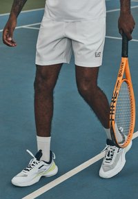 Calções de ténis brancos com um logótipo, combinados com sapatilhas de ténis brancas e azul-marinho com pormenores em verde. Jogador a segurar uma raquete de ténis laranja.