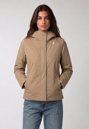 Vrouw met lang donker haar draagt een beige capuchonjas met een ritssluiting en zakken aan de voorkant, gecombineerd met een blauwe spijkerbroek, staand tegen een effen achtergrond.