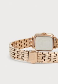 Montre en or rose avec un cadran carré, bracelet en maillons métalliques et fermoir. La montre présente une surface lisse avec un design minimaliste.