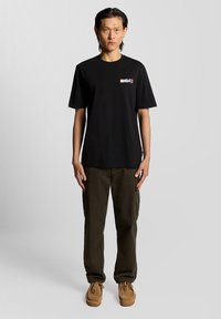 Melna kokvilnas t-krekls ar īsām piedurknēm, uz krūtīm ar daudzkrāsu grafiku, kopā ar olīvu krāsas kargo biksēm un brūnajām kurpēm.