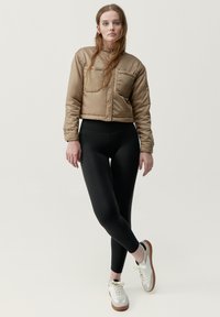 Korta, beige pufferjacka med en framficka, kombinerad med svarta leggings. Modellen har vita sneakers med bruna sulor och snören.