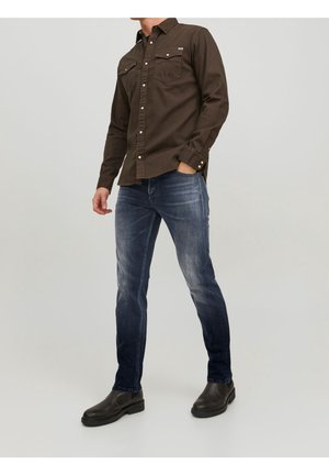 Ruskea pitkähihainen paita, jossa on kaksi rintataskua, yhdistettynä sinisiin farkkuihin, joissa on haalistunut kuviointi, ja mustiin nilkkureihin.